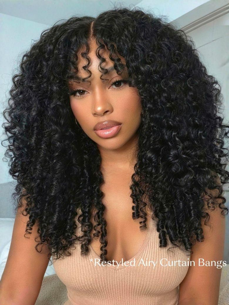 A woman with voluminous, defined curly hair and restyled wispy bangs poses indoors, showcasing a gorgeous Burmese curly wig. She wears a beige top and natural makeup.
